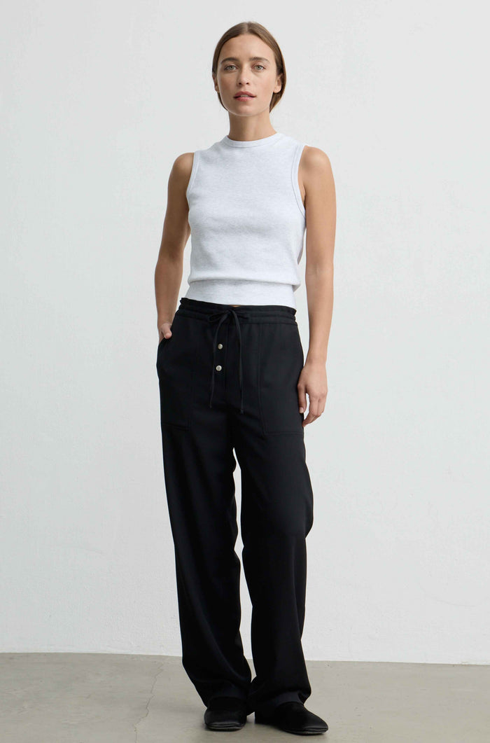 A woman stands against a plain light gray studio wall wearing a white sleeveless mock-neck top and loose black drawstring pants with front buttons.