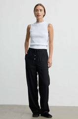 A woman stands against a plain light gray studio wall wearing a white sleeveless mock-neck top and loose black drawstring pants with front buttons.
