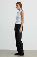 A woman stands in profile wearing a white sleeveless top and black wide-leg pants against a plain light gray studio background.