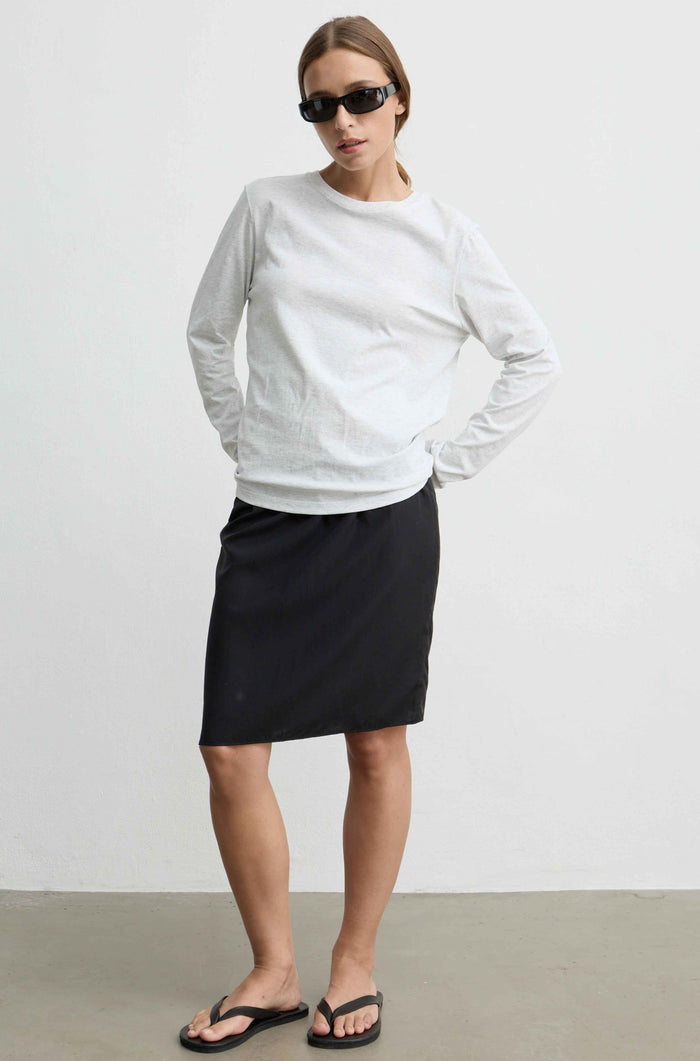 A woman modeling a white long-sleeve crewneck top and a black knee-length skirt, paired with black flip-flop sandals and sunglasses, standing against a plain light gray wall.