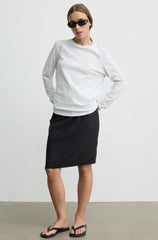 A woman modeling a white long-sleeve crewneck top and a black knee-length skirt, paired with black flip-flop sandals and sunglasses, standing against a plain light gray wall.