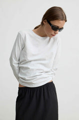 A woman wearing a light gray long-sleeve crewneck t-shirt and black pants, photographed against a plain light gray background.