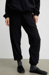 A person wearing loose black jogger-style pants with a drawstring waist, paired with gray socks and black slip-on shoes, photographed against a plain light wall and concrete floor.