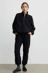 A woman modeling a black fleece half-zip pullover and matching black fleece jogger pants against a plain light gray studio background.