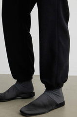 Close-up of a person's lower legs wearing black jogger sweatpants, dark gray ribbed socks, and black slip-on shoes on a light gray floor against a white wall.