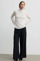 A woman modeling a cream turtleneck sweater and black wide-leg trousers against a plain light gray studio background.