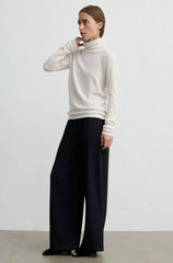 A woman modeling a cream-colored turtleneck sweater and black wide-leg trousers against a plain light gray studio background.