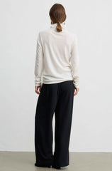 A woman viewed from behind wearing a cream-colored turtleneck sweater and black wide-leg trousers, standing against a plain white wall.