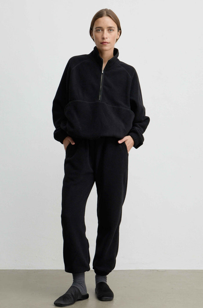 A woman modeling a black fleece half-zip pullover and matching black fleece jogger pants against a plain light gray studio background.