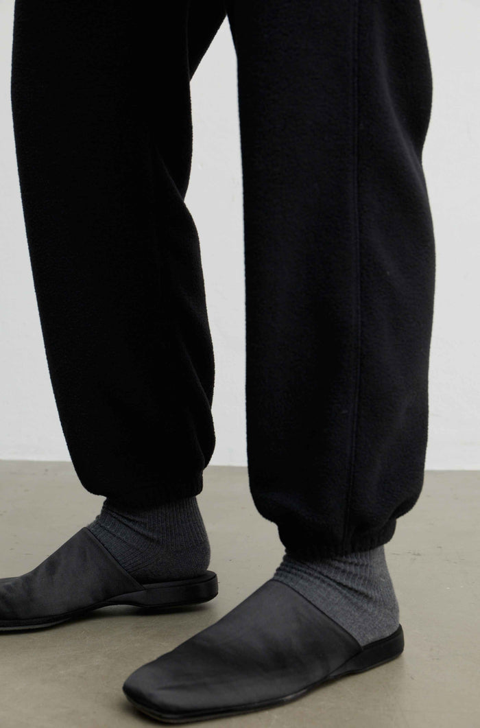 Close-up of a person's lower legs wearing black jogger sweatpants, dark gray ribbed socks, and black slip-on shoes on a light gray floor against a white wall.