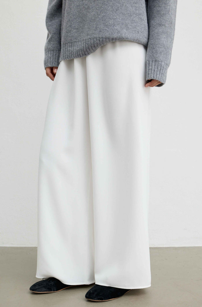 A person wearing wide-leg white trousers, a gray knit sweater, and black slippers against a plain light gray wall and concrete floor.