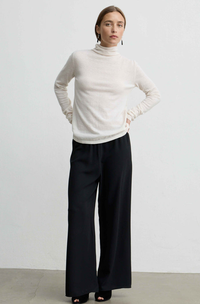 A woman modeling a cream turtleneck sweater and black wide-leg trousers against a plain light gray studio background.