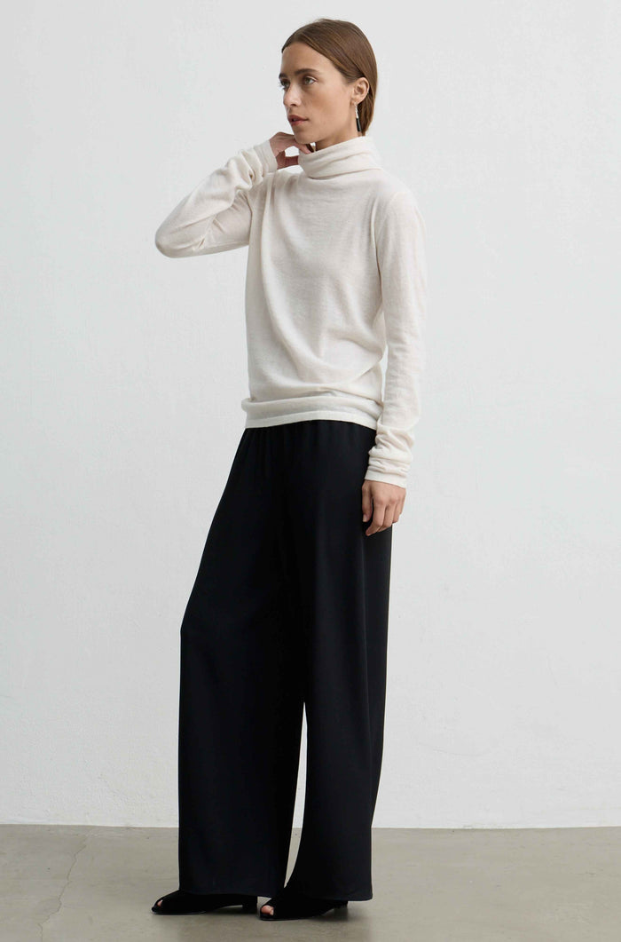 A woman modeling a cream-colored turtleneck sweater and black wide-leg trousers against a plain light gray studio background.