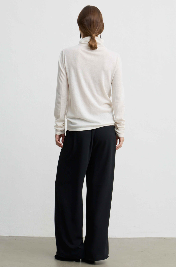 A woman viewed from behind wearing a cream-colored turtleneck sweater and black wide-leg trousers, standing against a plain white wall.