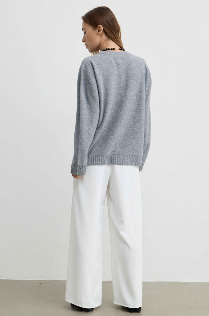 A woman shown from the back wearing a light gray knit sweater and white wide-leg trousers, standing against a plain light gray studio wall.